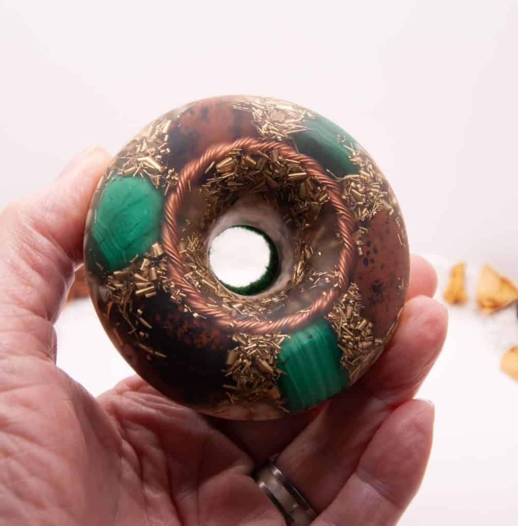 Handheld view of a torus shaped resin case filled with Malachite, Mahogany Obsidian, Quartz and Dolomite crystals. Copper ring and Brass shavings are also inside. The backing is Green Felt